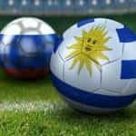 A soccer ball with the Uruguayan flag on it rests on a soccer field in front of a second soccer ball with the Russian flag on it.