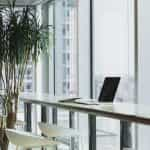 A laptop sitting at an empty workspace in a corporate office facing large floor to ceiling windows.